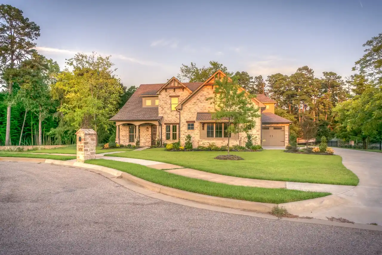 expansive contemporary luxury home with floor-to-ceiling glass, natural stone exterior, landscaped driveway, and warm evening lighting on a wooded property
