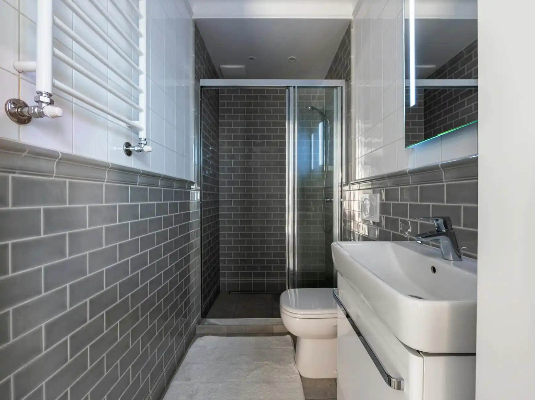 compact bathroom remodel with light oak vanity, matte black faucet, white subway tile shower surround, hex floor tile, and warm layered lighting