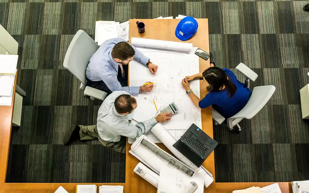 architect and homeowners reviewing custom house blueprints at a large wood table, modern mountain home elevations, material samples, and natural daylight from floor-to-ceiling w...