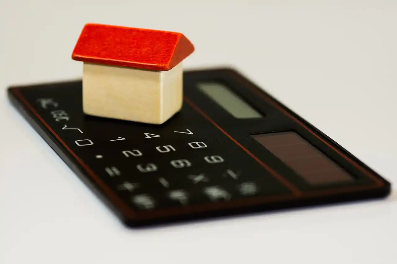 close-up of a lender and homebuyers reviewing custom home blueprints at a bright office table, with calculator, coffee mugs, and a tablet showing mortgage numbers