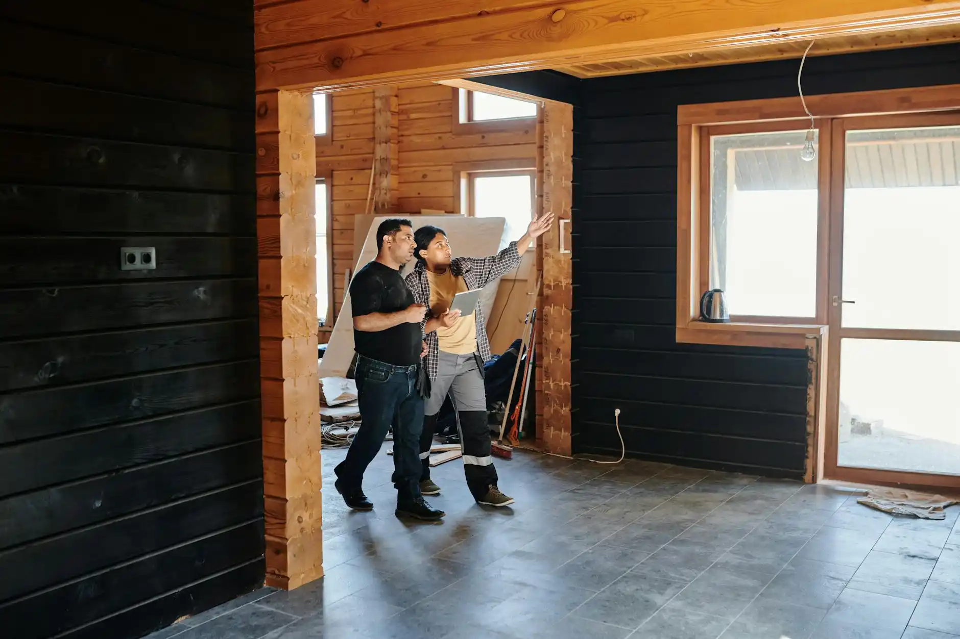 contractor in branded workwear walking through a partially renovated home with clipboard, measuring tape, and lender renovation checklist, exposed framing and new windows visible
