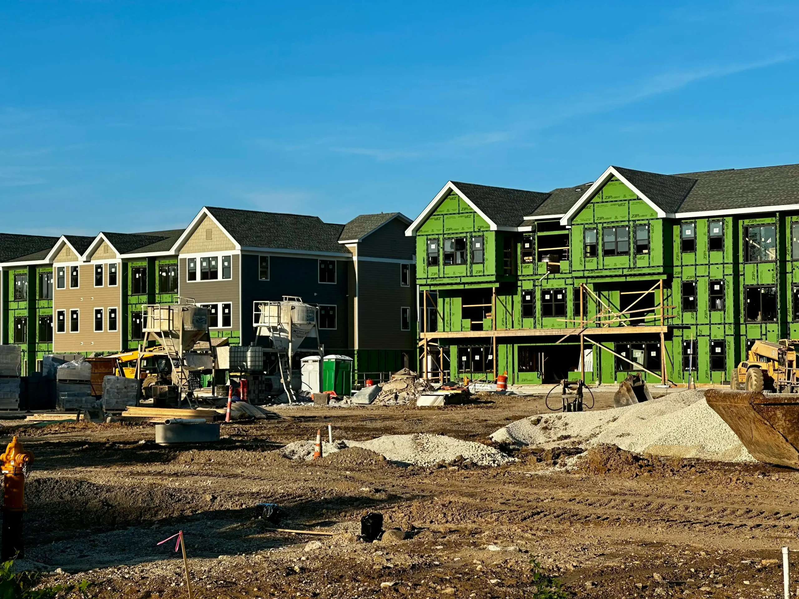 Seattle residential construction site with excavator on a steep lot, retaining wall forms, orange safety fencing, and neighboring craftsman homes