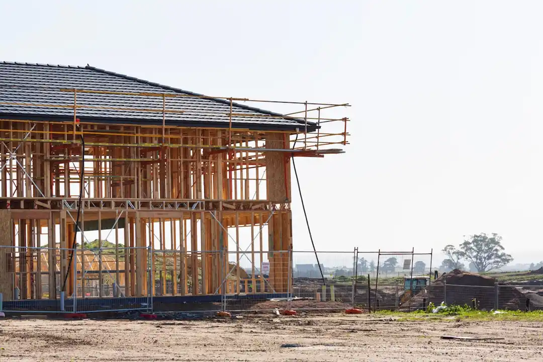 luxury custom home under construction with framed exterior, crane lifting roof trusses, and a wooded suburban lot at sunrise