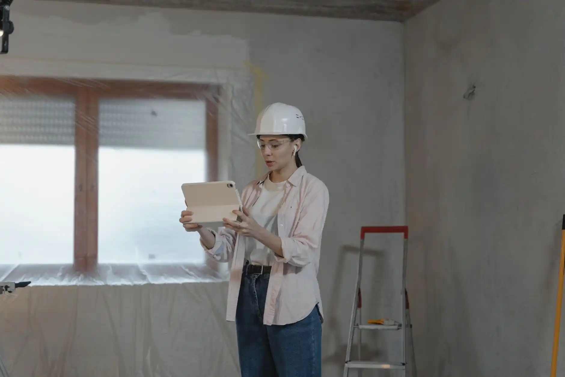 residential construction site with framing underway, superintendent in hard hat reviewing progress on a tablet beside stacks of lumber and partially installed windows